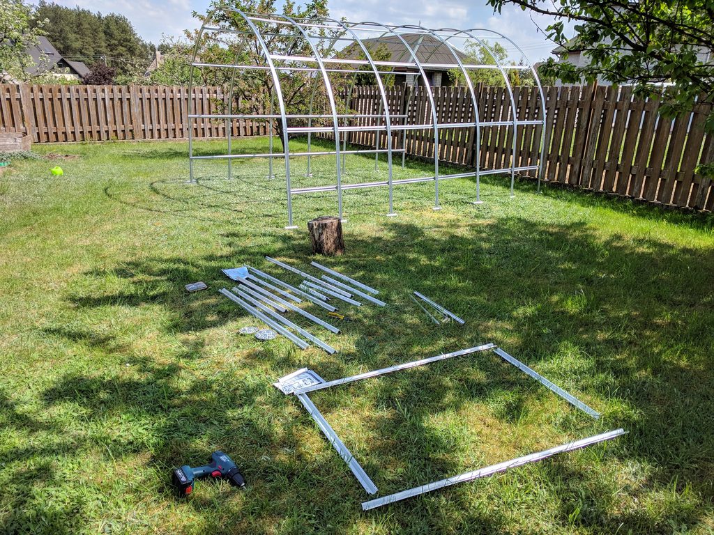 Arched metal greenhouse frame on the lawn, with loose components and a cordless drill nearby.

